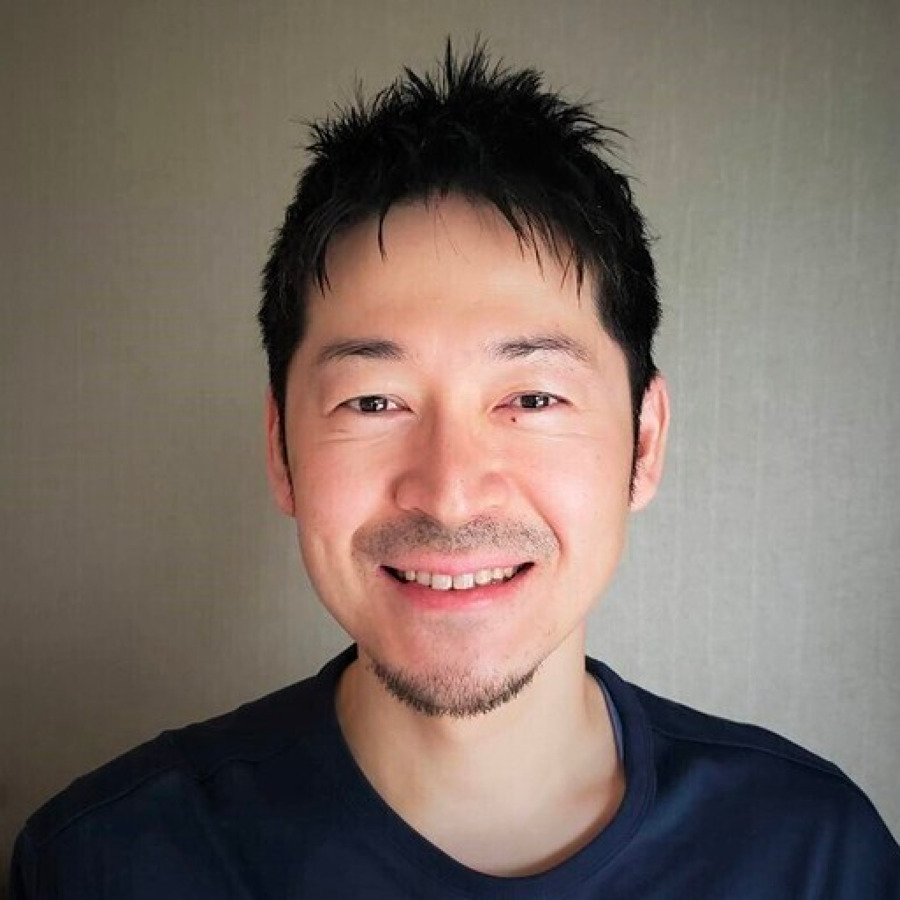 Portrait of a man with short dark hair, smiling at the camera, wearing a black shirt, in an indoor setting with a soft-focus background.