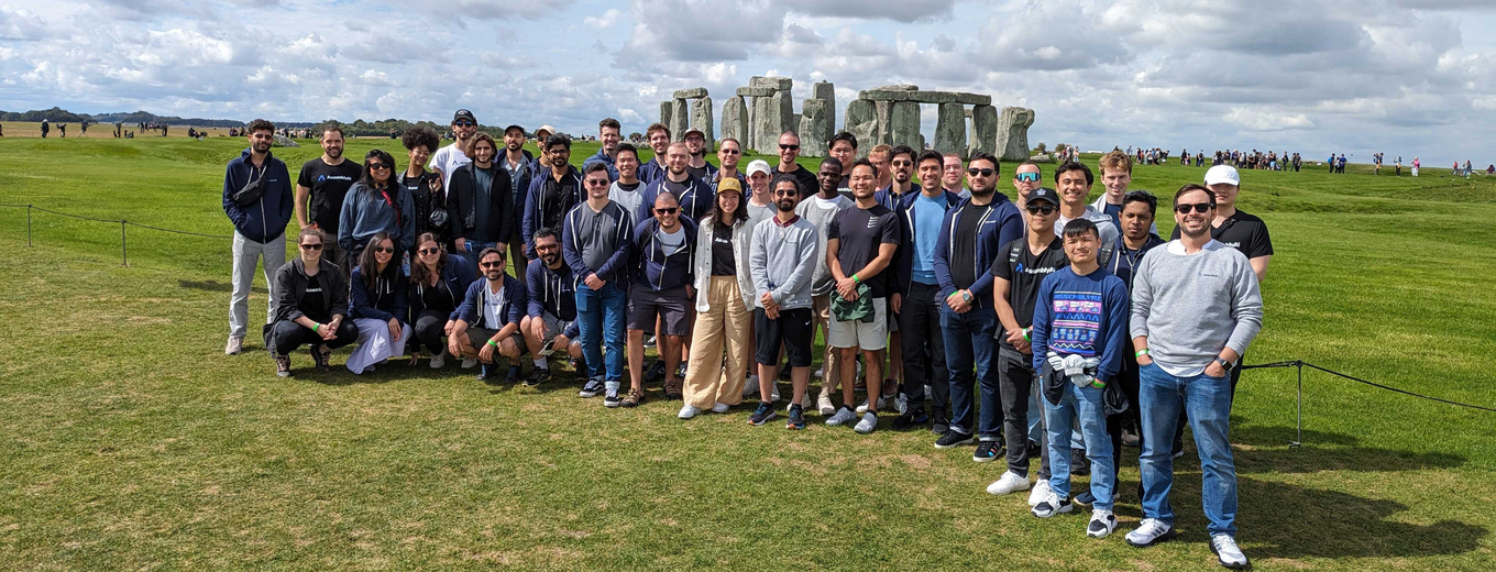 Group photo of diverse individuals in casual clothing, posing in front of the Stonehenge under a cloudy sky. Some are standing while others are kneeling on the grass.